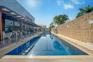 A modern, rectangular outdoor swimming pool with clear blue water stretches alongside a building with large glass windows. The pool area has sun loungers and tropical plants, with a stone wall on one side and a sunny, blue sky overhead.