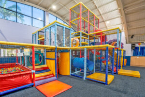 Indoor play area with colorful climbing structures, slides, and obstacle courses enclosed in netting. The room is well-lit with large windows that let in natural light, and there is a ball pit on the left side. The flooring is padded for safety, and the ceiling is high with exposed beams pointing like a North Star to fun.