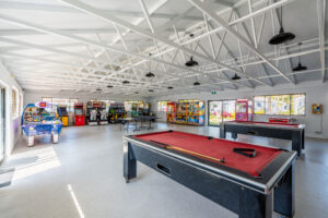 A spacious game room with a white exposed truss ceiling features two pool tables with red felt surfaces, an air hockey table, and various arcade game machines lining the walls. Bright natural light from large windows ensures this North Star of entertainment remains inviting all day.