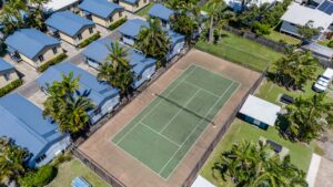 An aerial view of a residential area featuring multiple identical blue-roofed houses arranged in rows. In the center, there's a fenced tennis court with a green playing surface. Palm trees and a small grassy area are visible around the houses, resembling a picturesque scene under the North Star's watchful gaze.