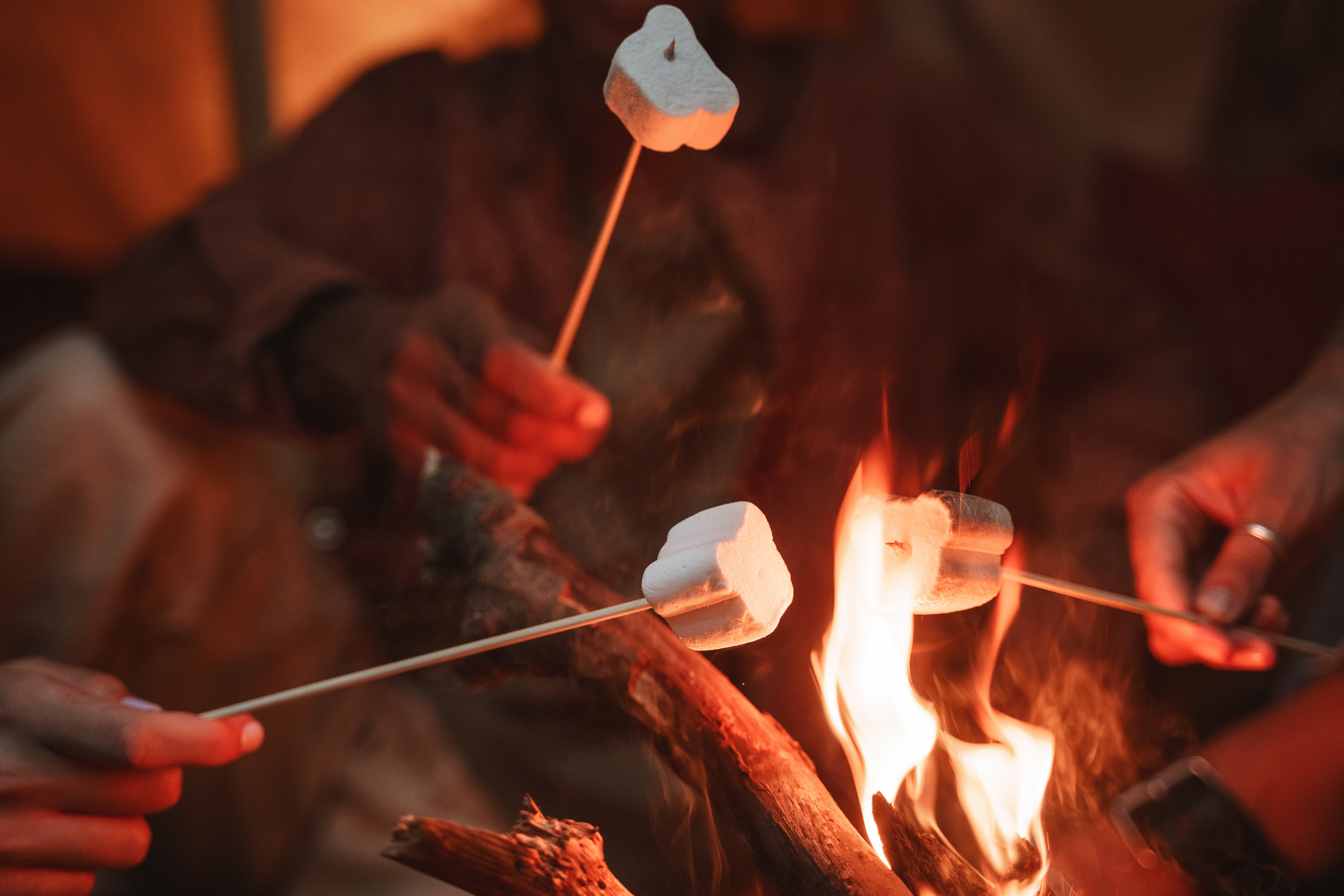 Three people roast marshmallows on sticks over a campfire. The flames flicker and illuminate the marshmallows, which are held at different angles above the fire. The scene has a warm, cozy ambiance, suggesting a camping or outdoor gathering at night.