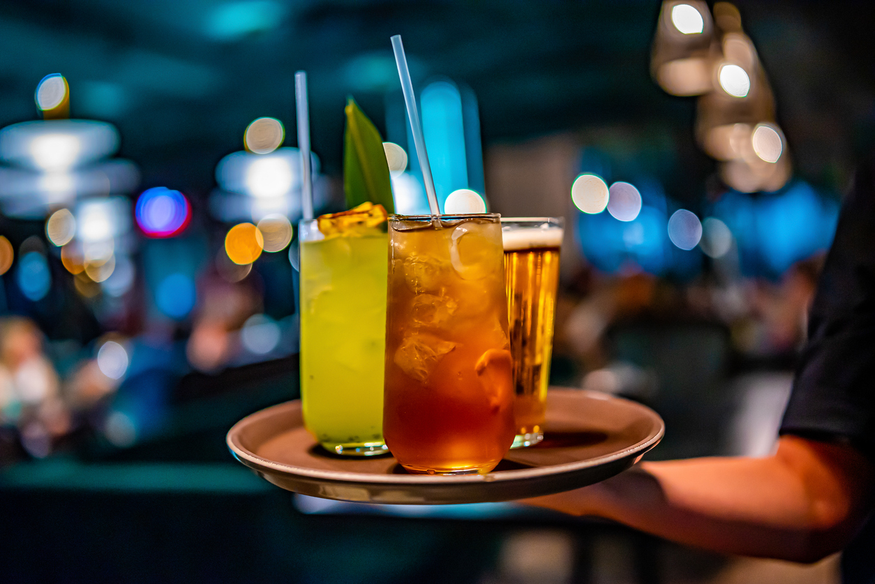 A close-up view of a server's hand holding a tray with three drinks: a green cocktail with a leaf garnish, an amber-colored drink filled with ice, and a glass of beer. The background is blurred, featuring bokeh lights in a dimly lit bar or restaurant.