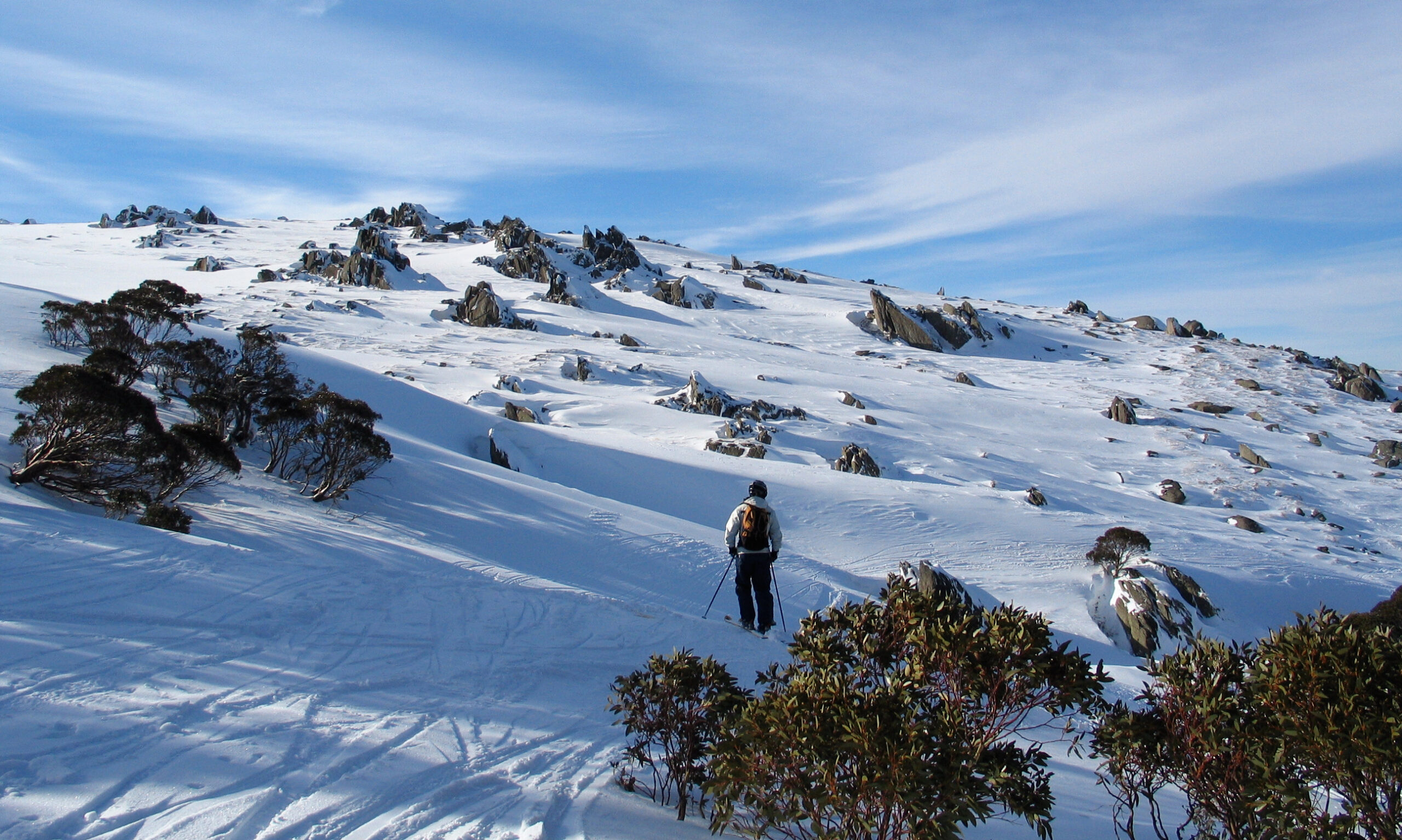 A lone skier in a blue jacket and black backpack traverses a snowy mountain slope dotted with rocks and sparse bushes. Tracks in the snow indicate prior skier activity. The sky above is mostly clear with some wispy clouds.