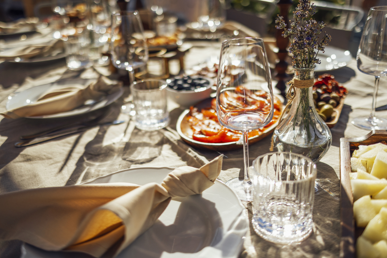 A sunlit table set for a meal, featuring plates with beige napkins, empty wine glasses, and various dishes, including sliced tomatoes, blueberries, and olives. A vase of lavender flowers is placed among the tableware, creating an elegant dining ambiance.