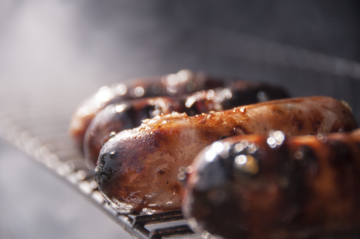 Close-up of several grilled sausages with a smoky background. The sausages are charred in places, showing a glossy, well-cooked appearance. They are placed on a metal rack, likely over a barbecue grill. Steam or smoke is visible, enhancing the freshly grilled effect.