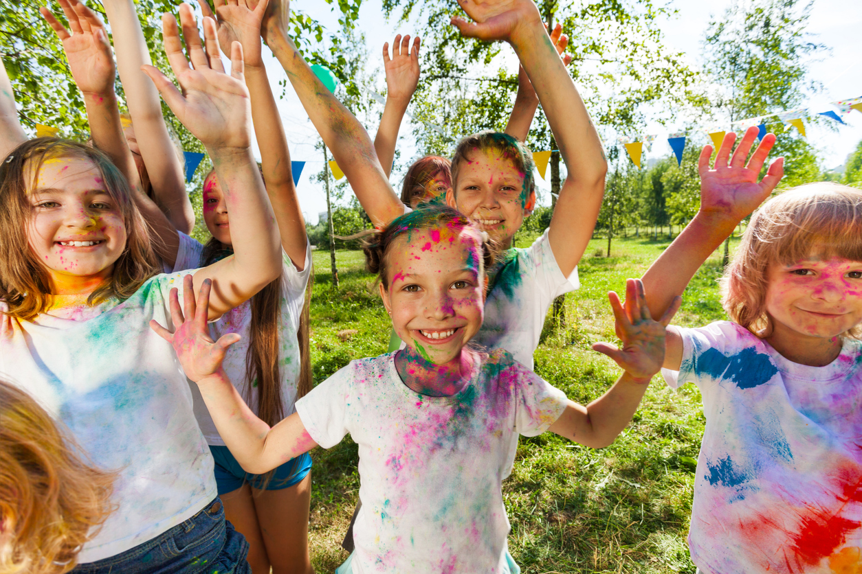 A group of children with colorful powder on their faces and clothes are raising their hands and smiling joyfully. They are standing outdoors on a sunny day with trees and bunting flags in the background. Everyone appears to be enjoying a festive, playful event.