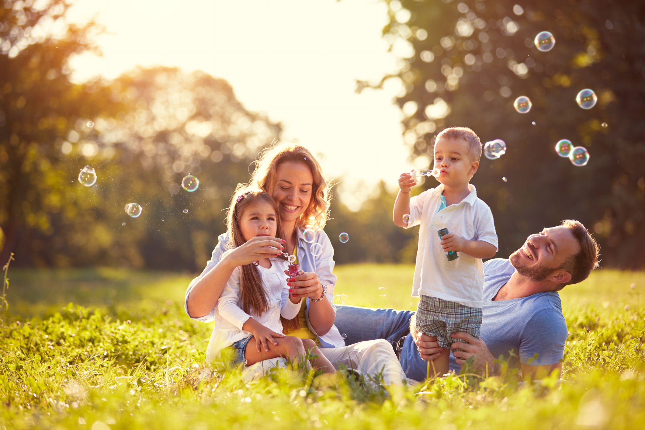 A family of four enjoys a sunny day in a meadow. The parents sit on the grass, smiling, as the young girl sits on her mother's lap, and the boy stands nearby. They are all blowing bubbles, with some bubbles floating around them. Trees and sunlight fill the background.