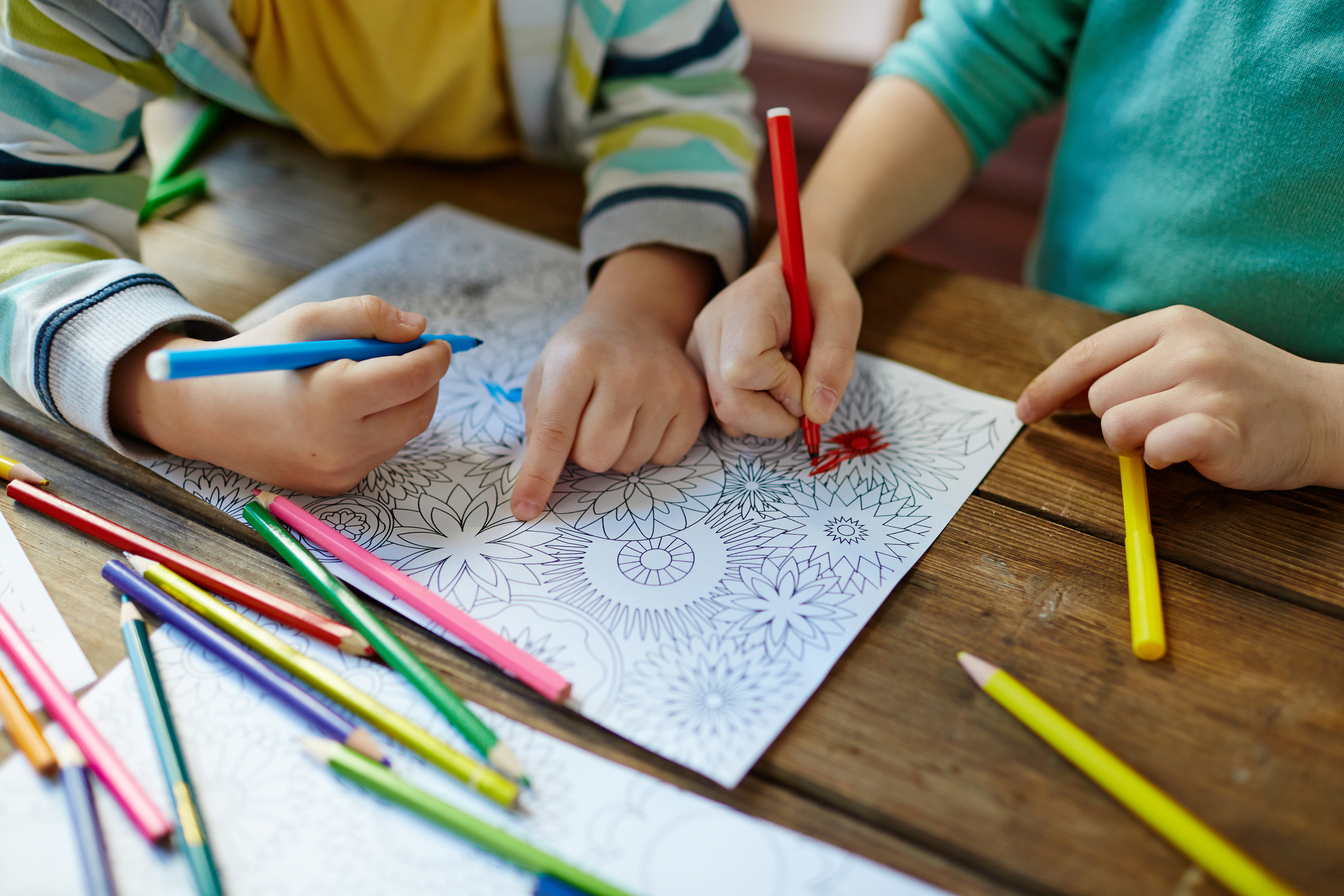 Two children are coloring an intricate floral coloring page with colored pencils at a wooden table. One child uses a blue pencil while the other uses a red pencil. Various other colored pencils are scattered around the table.