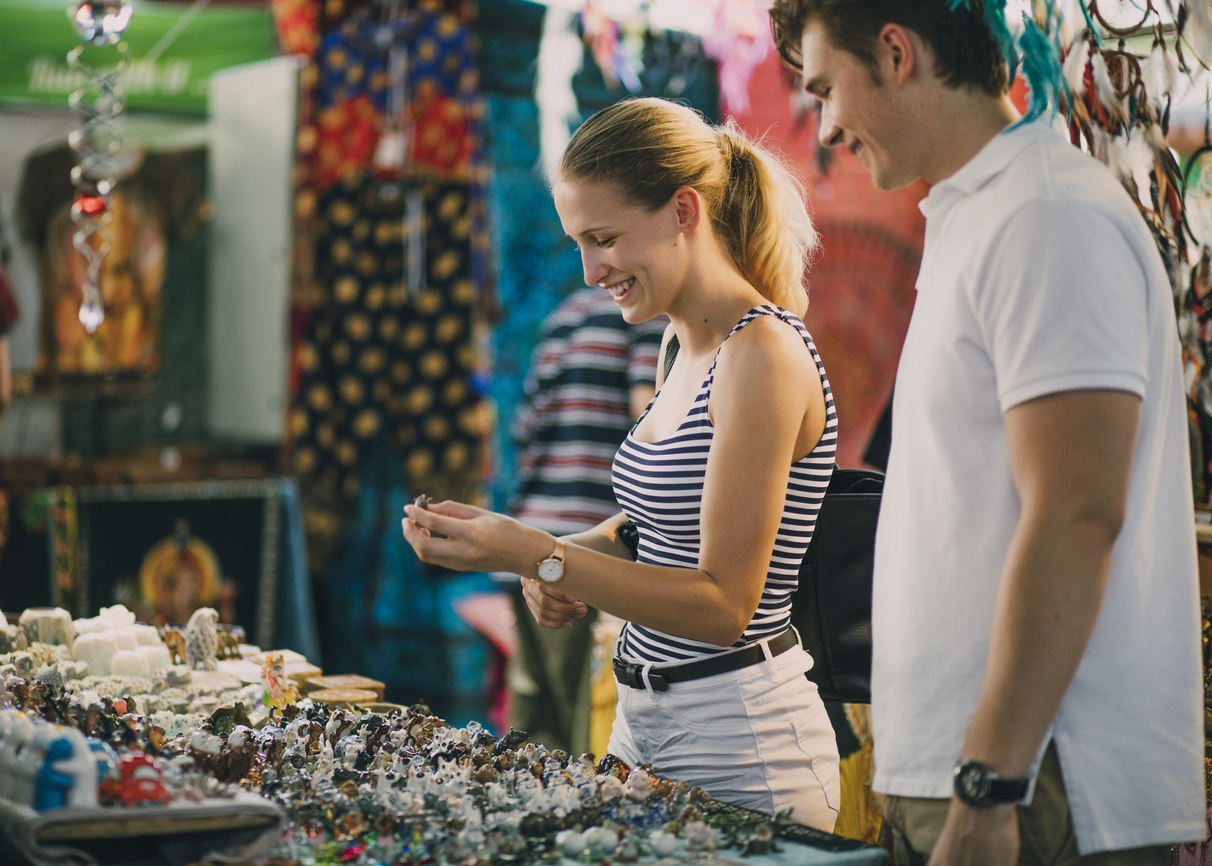 A young woman and man happily browse a colorful outdoor market stall filled with various trinkets and souvenirs. The woman is holding an item in her hand, while the man stands beside her, both smiling and enjoying the experience.