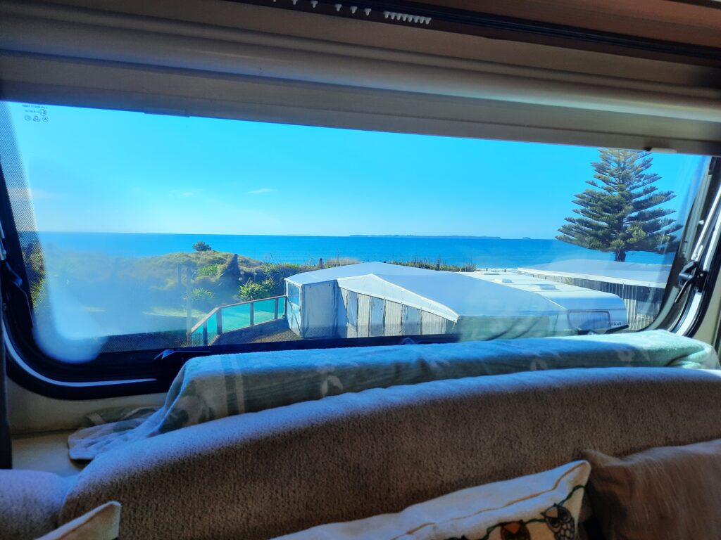 A scenic view from inside a camper van, showing a clear blue ocean and a pine tree through the window. Green grass and white-roofed structures are visible outside, with a sofa and cushions in the foreground. It appears to be a sunny day.