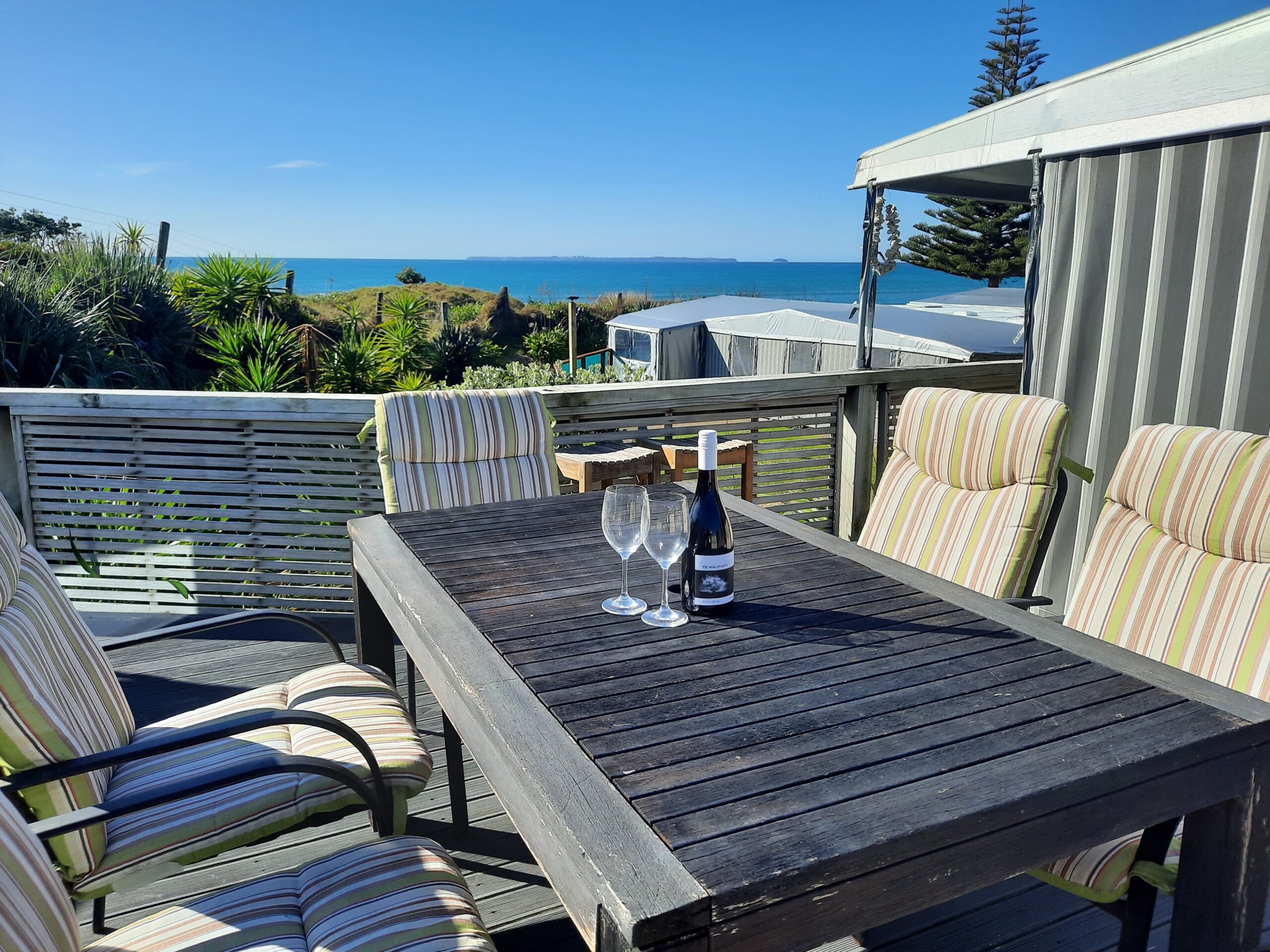 A seaside deck with a wooden table and striped cushioned chairs. On the table, there are two wine glasses and a bottle of wine. The deck overlooks a vibrant blue ocean under a clear sky. Various green plants surround the area, adding to the tranquil setting.