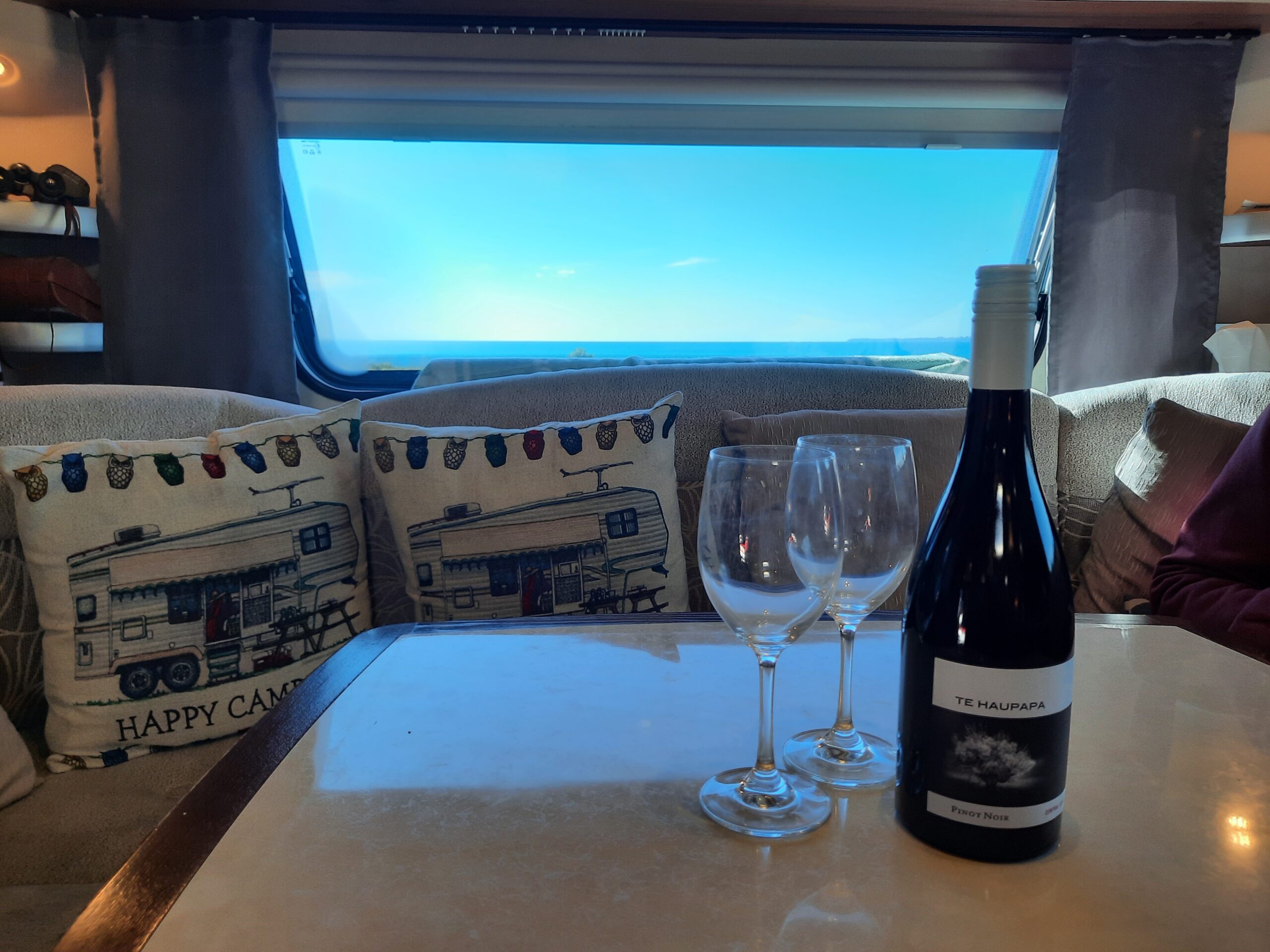 A cozy RV interior with a large window revealing a scenic landscape. The table is set with a bottle of red wine and two empty wine glasses. The couch displays patterned pillows featuring a camper design and the phrase "Happy Camper.