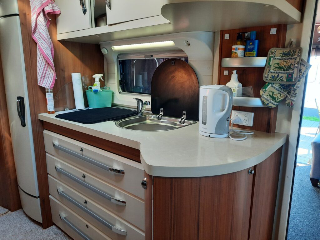 Compact kitchen area with light wood cabinetry, featuring a small sink, electric kettle, and various cleaning supplies. There's a counter with multiple drawers below, a mounted paper towel holder, and a window above the sink with a blind partially pulled down.