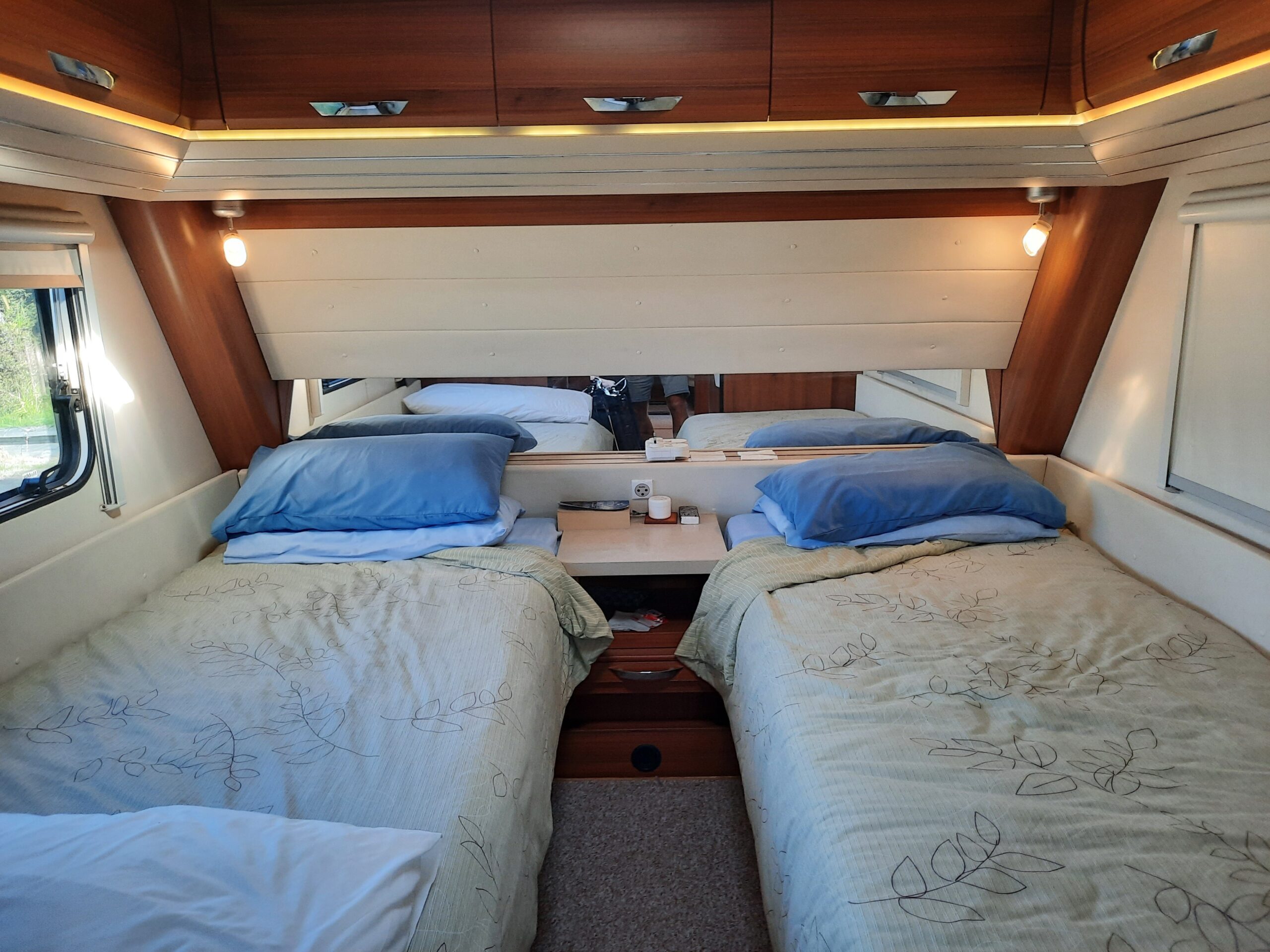 Interior of a cozy, well-lit RV bedroom with two single beds placed parallel to each other. Each bed has blue pillows and patterned beige bedspreads. There are overhead cabinets, wall-mounted lights, and a small central nightstand with books and a tissue box.