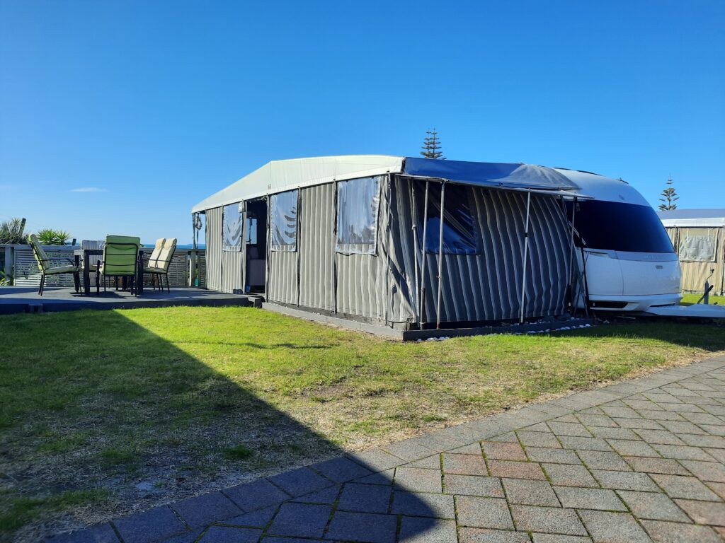 A caravan is covered with a striped, protective awning at a campsite on a sunny day. The setup includes a paved patio area with a table and several outdoor chairs to the left. The sky is clear and blue, with some greenery visible in the background.