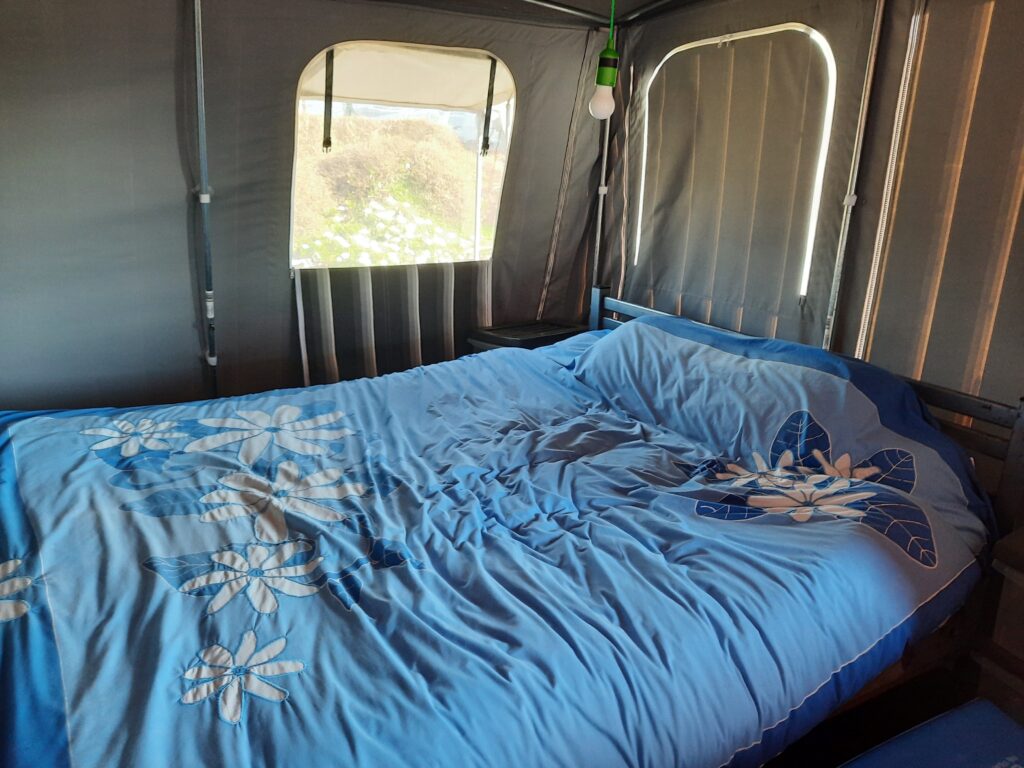 A cozy bed with blue floral bedding inside a tent. The tent has large windows with clear panels, letting in natural light. A hanging light bulb is visible above the bed, providing additional illumination. The setting suggests an indoor camping setup.