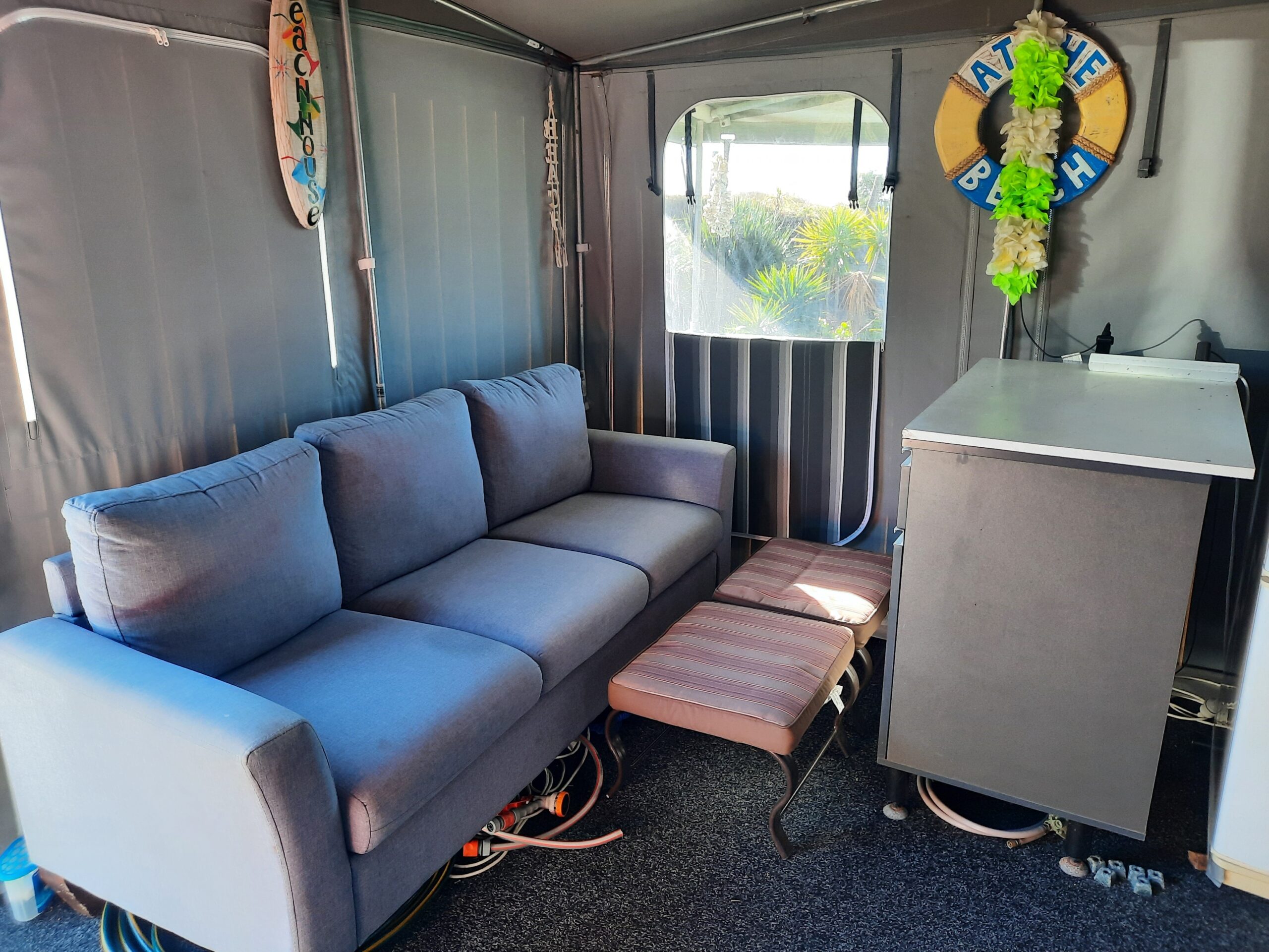 A cozy indoor space with a gray sofa, two striped ottomans, and a small counter. Decorations include surfboards, a lifebuoy with "The Beach" and green flowers, and various beach-themed accessories. The area has gray walls and carpeting.