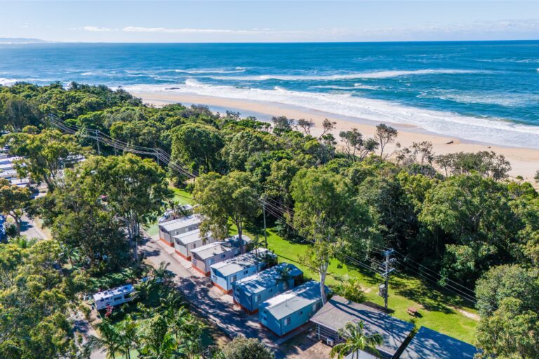 A scenic aerial view of a coastal area with a row of cabins near a beach, surrounded by lush green trees and vegetation, now kissed by winter's frost. The clear blue ocean and waves shimmer in the background under a partly cloudy sky.