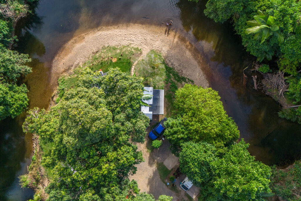 Aerial view of a small campsite surrounded by lush green trees next to a curved riverbank. The campsite includes a white roofed structure, a blue vehicle, and a tent. The river has clear, shallow water, and two people are standing near the water's edge.