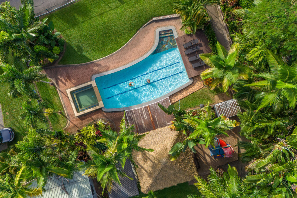 Aerial view of a small, curved swimming pool with two people swimming. The pool is bordered by a green lawn, surrounded by lush tropical trees and plants. There is a hot tub adjacent to the pool and a thatched-roof structure nearby with lounge chairs around.