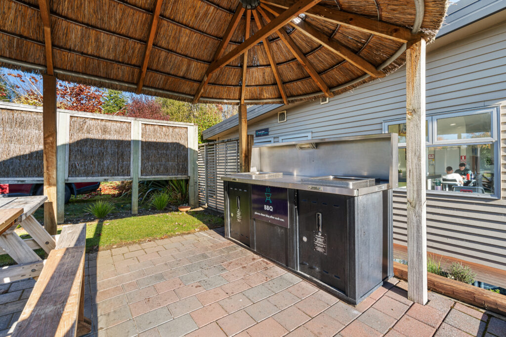 Outdoor BBQ area with a thatched roof gazebo. The stainless steel grilling station is set on a brick patio and surrounded by a wooden fence. A nearby building has large windows. A wooden picnic bench is also visible.