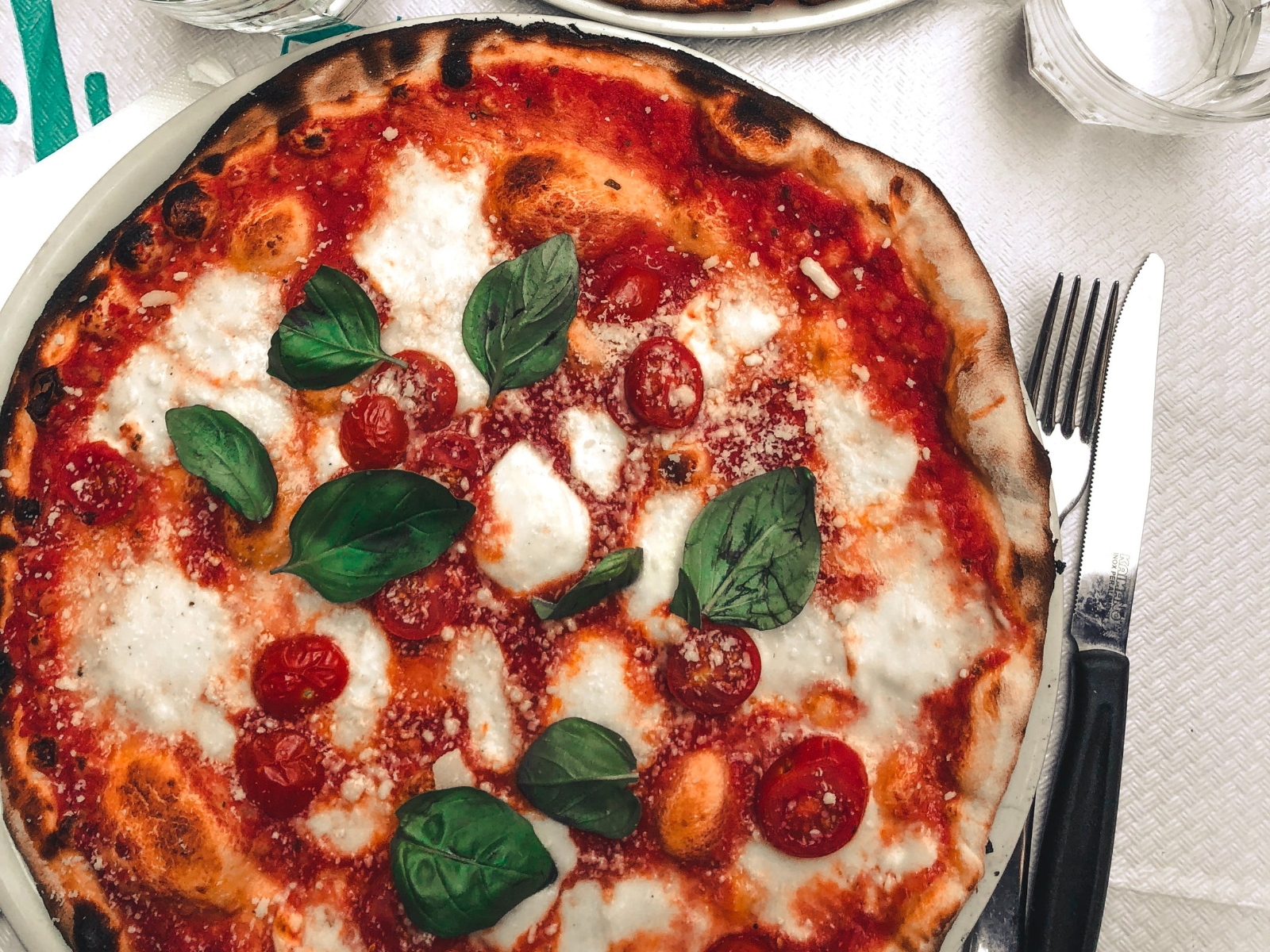 A close-up of a freshly baked pizza topped with melted mozzarella cheese, vibrant tomato sauce, whole cherry tomatoes, and fresh basil leaves. The pizza has a slightly charred, crispy crust. A knife and fork are placed to the right of the plate on a white tablecloth.