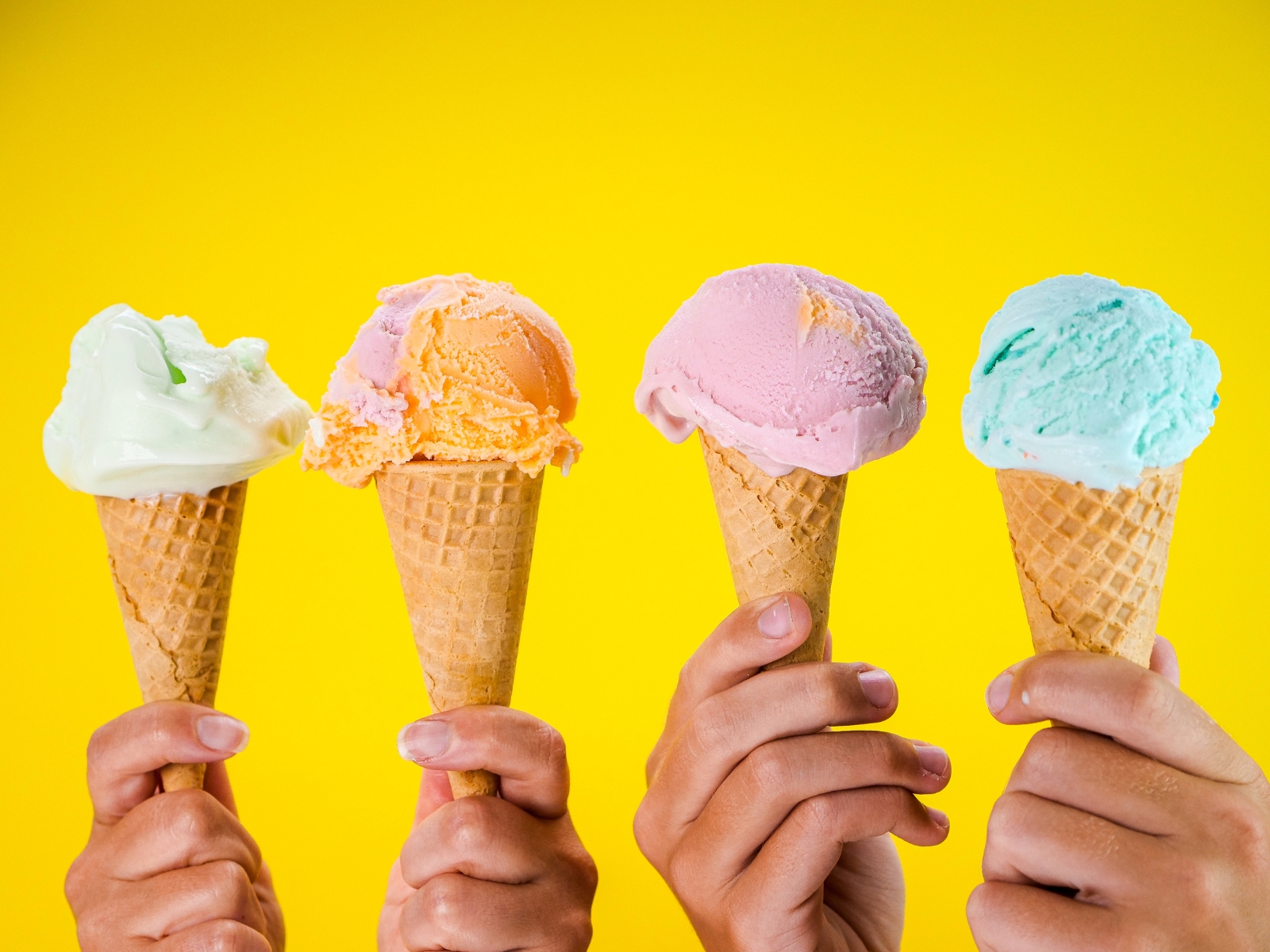 Four hands hold up ice cream cones against a bright yellow background. The ice cream flavors, from left to right, are green, orange, pink, and blue. Each scoop is perched on a waffle cone.