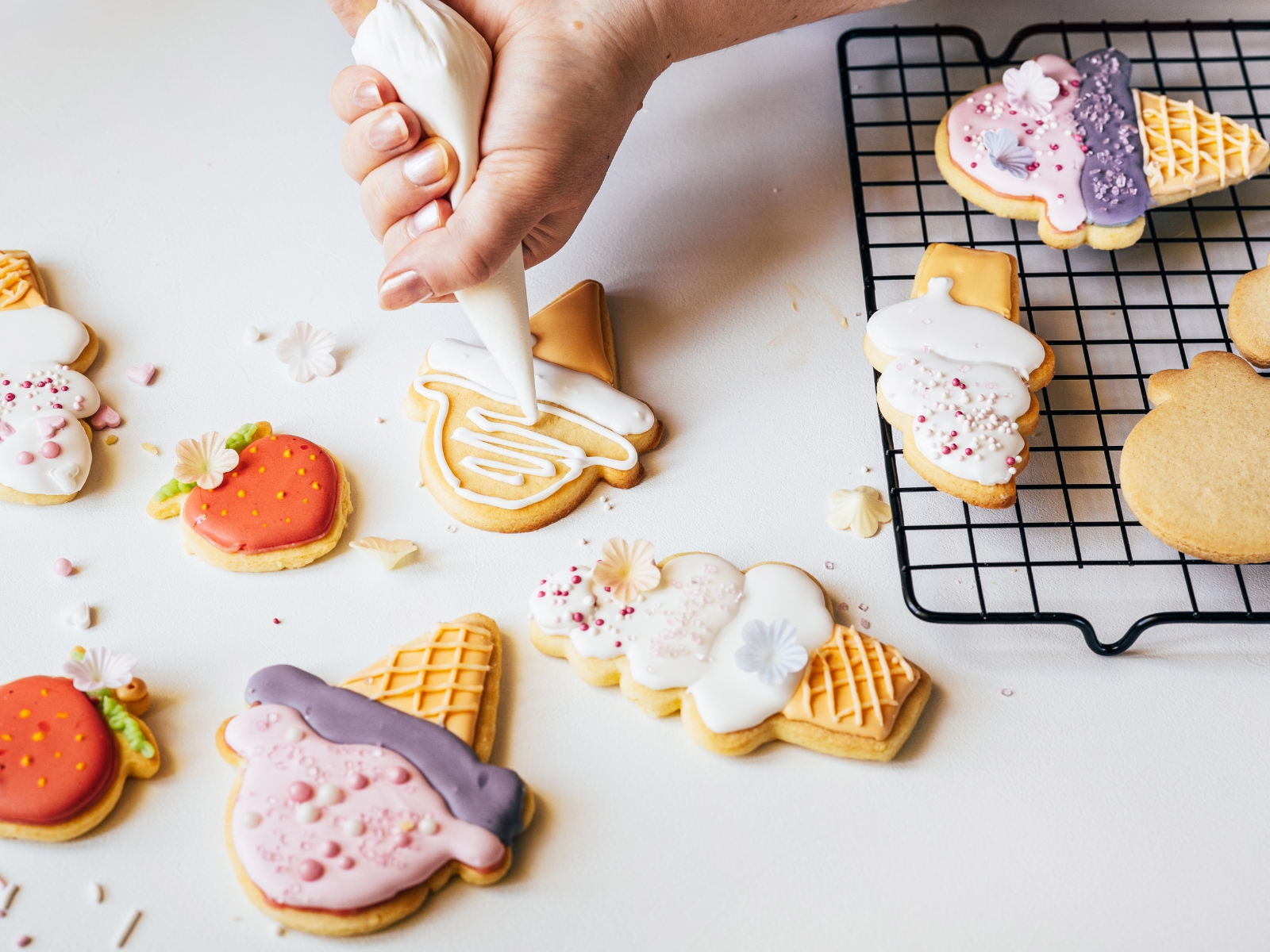 A person's hand is piping white icing onto a sugar cookie shaped like an ice cream cone. Other decorated cookies, resembling ice cream cones and popsicles, are on a cooling rack and scattered on a white surface. Some sprinkles and small edible decorations are also visible.