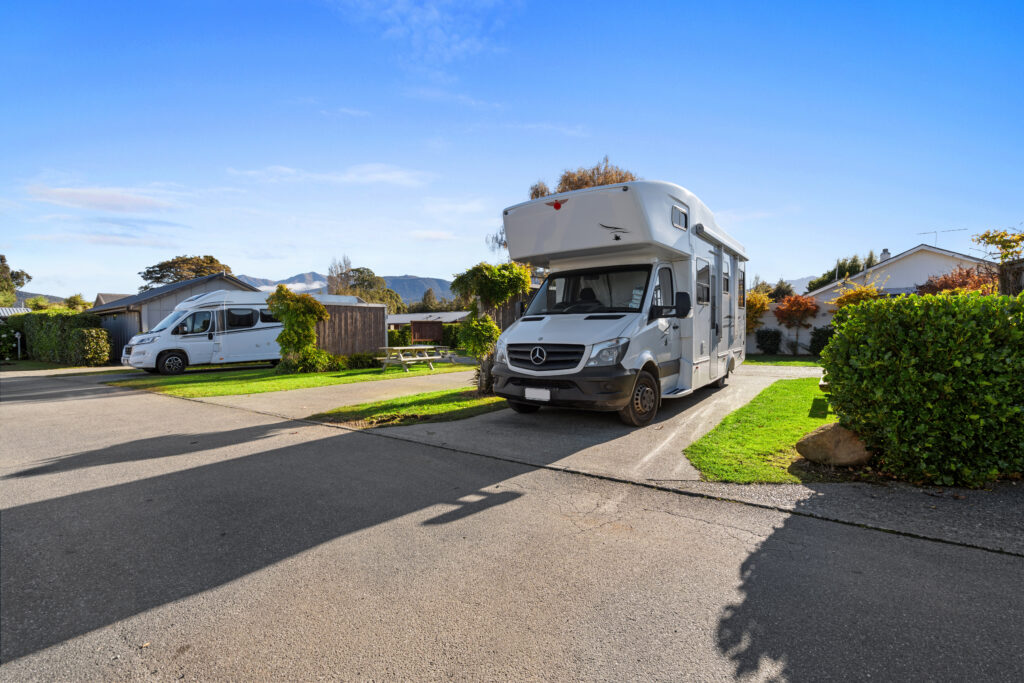 Two motorhomes are parked on neatly maintained driveways in a residential neighborhood surrounded by greenery. The sky is bright and blue, with distant mountains in the background. One motorhome is in the foreground, and the other is further down the road.