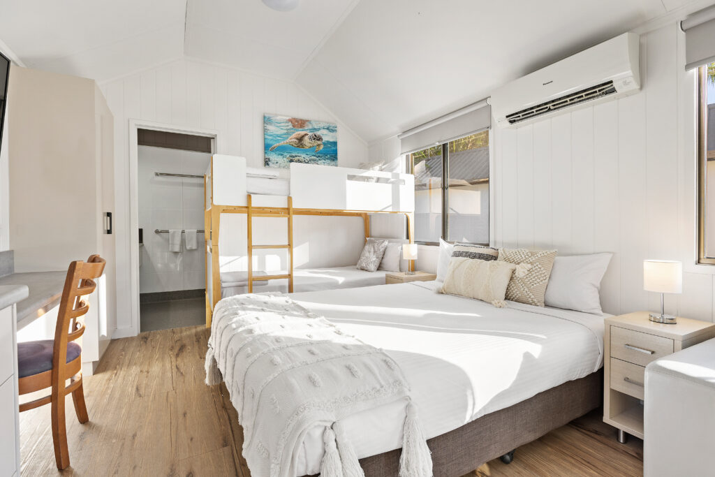 A bright, modern bedroom with wooden flooring, featuring a double bed adorned with white linens and decorative pillows. Above and to the side of the bed are bunk beds. Natural light floods through the windows, and an air conditioner is mounted on the white wall.