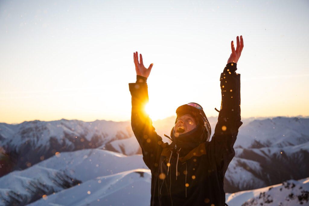 Person in winter clothing stands with arms raised, surrounded by glistening particles against a stunning backdrop of snow-covered mountains and a setting sun. The scene captures the joy and majesty of a winter adventure.