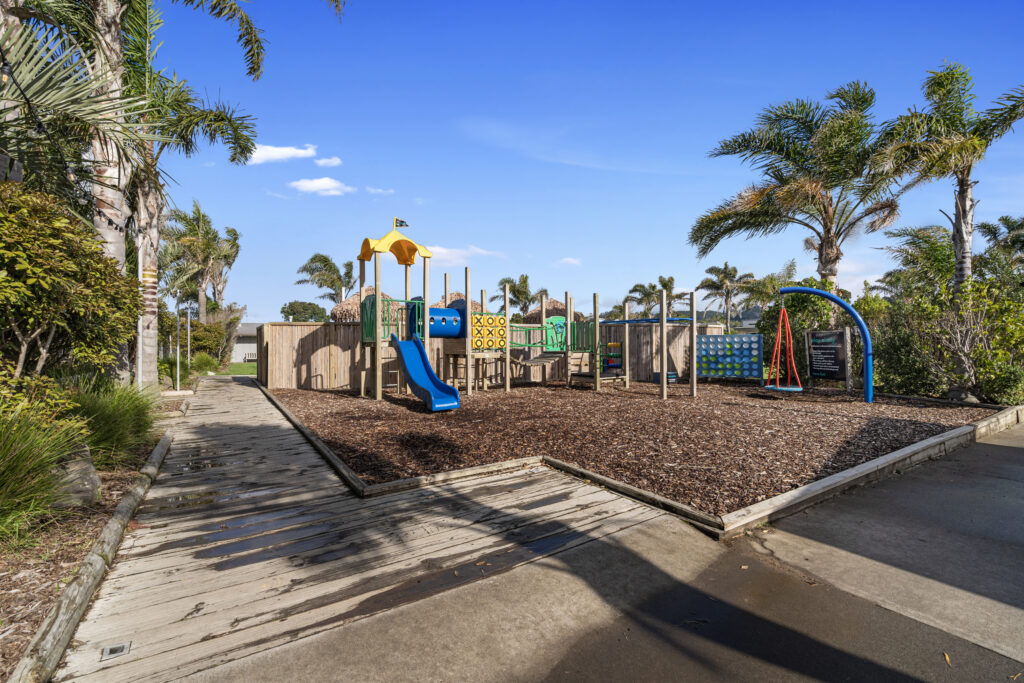 A playground with various equipment, including a slide, tic-tac-toe board, climbing structures, and swings, surrounded by wood chips. It is set against clear blue skies and palm trees, with a wooden walkway and concrete path adjacent. Perfect for school holiday fun!