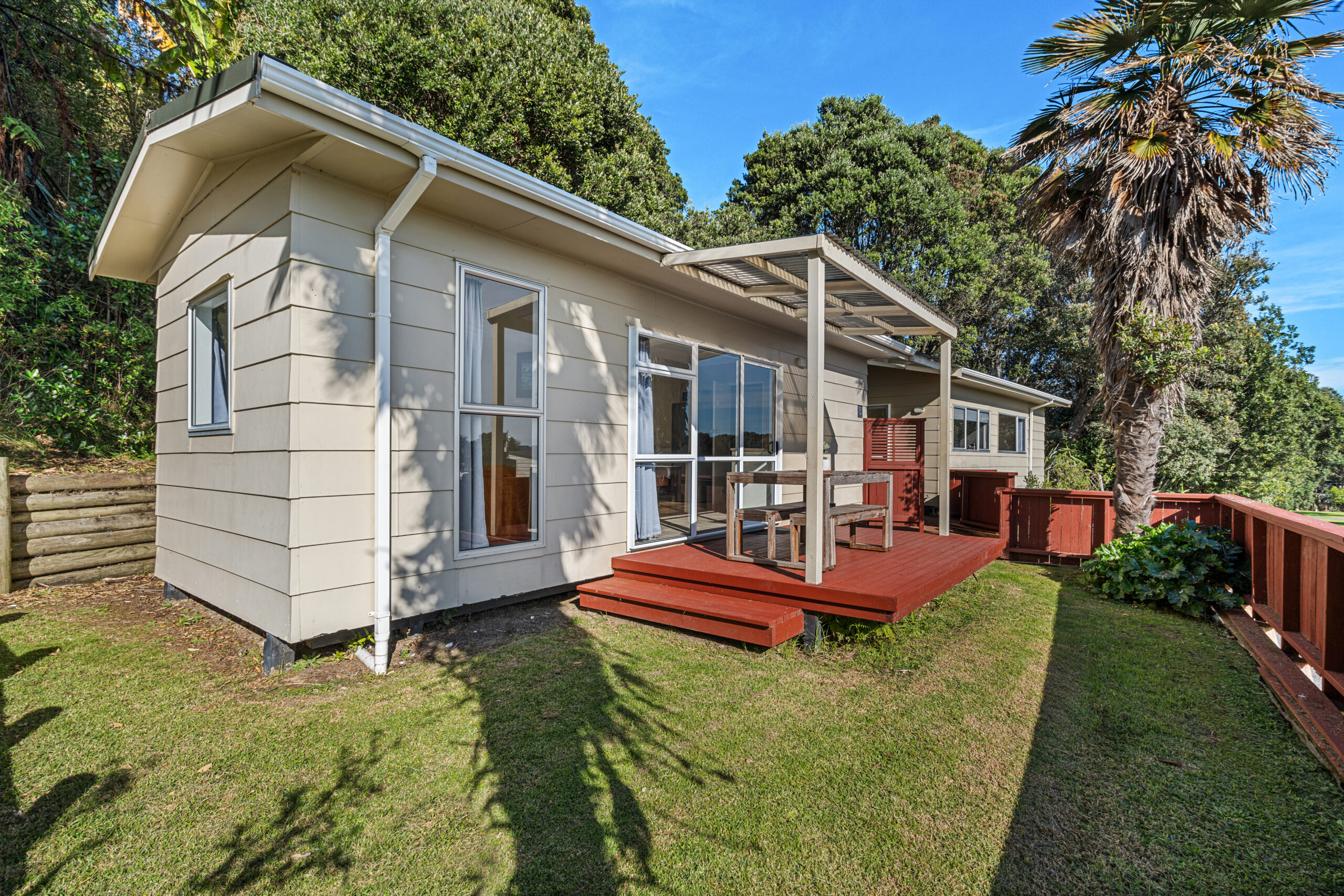 A cream-colored house with a wooden deck, surrounded by green lawn and lush trees. The deck features outdoor seating and a partial pergola. A palm tree is visible in the foreground, and the sky is clear with bright blue color.