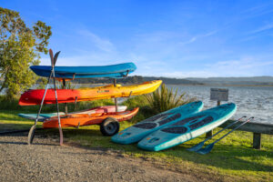 A sunny beachside scene featuring a rack holding three brightly colored kayaks and paddles on a gravel path. Two turquoise paddleboards with paddles rest on the grass nearby. Trees, reeds, and distant hills line the opposite shore under a clear blue sky.