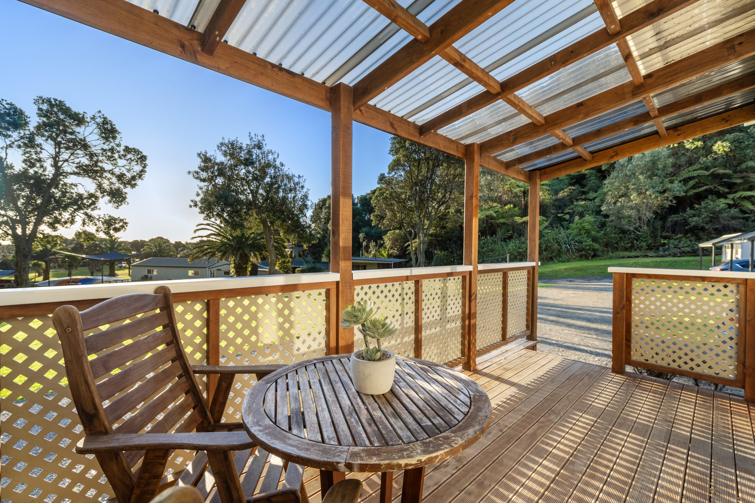 The image shows a wooden deck with a small round table, two wooden chairs, and a potted succulent plant. The deck is covered with a clear roof and overlooks a scenic outdoor area with trees and a gravel pathway, under a clear blue sky.