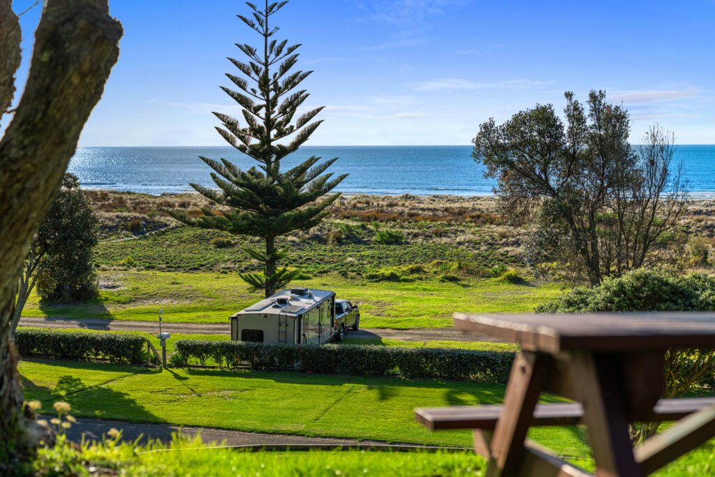 A motorhome is parked near a scenic coastal area with a clear view of the ocean. Tall trees and a manicured lawn surround the area, with a picnic table in the foreground. The sky is clear and blue, enhancing the serene atmosphere of the landscape.