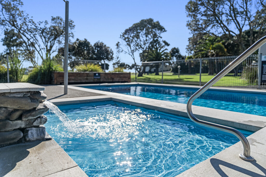 A bright, clear day at an outdoor swimming pool with crystal blue water. The pool features a waterfall spout and a metal handrail. Trees and a grassy area are in the background, surrounded by a tall fence. The pool area appears clean and inviting.