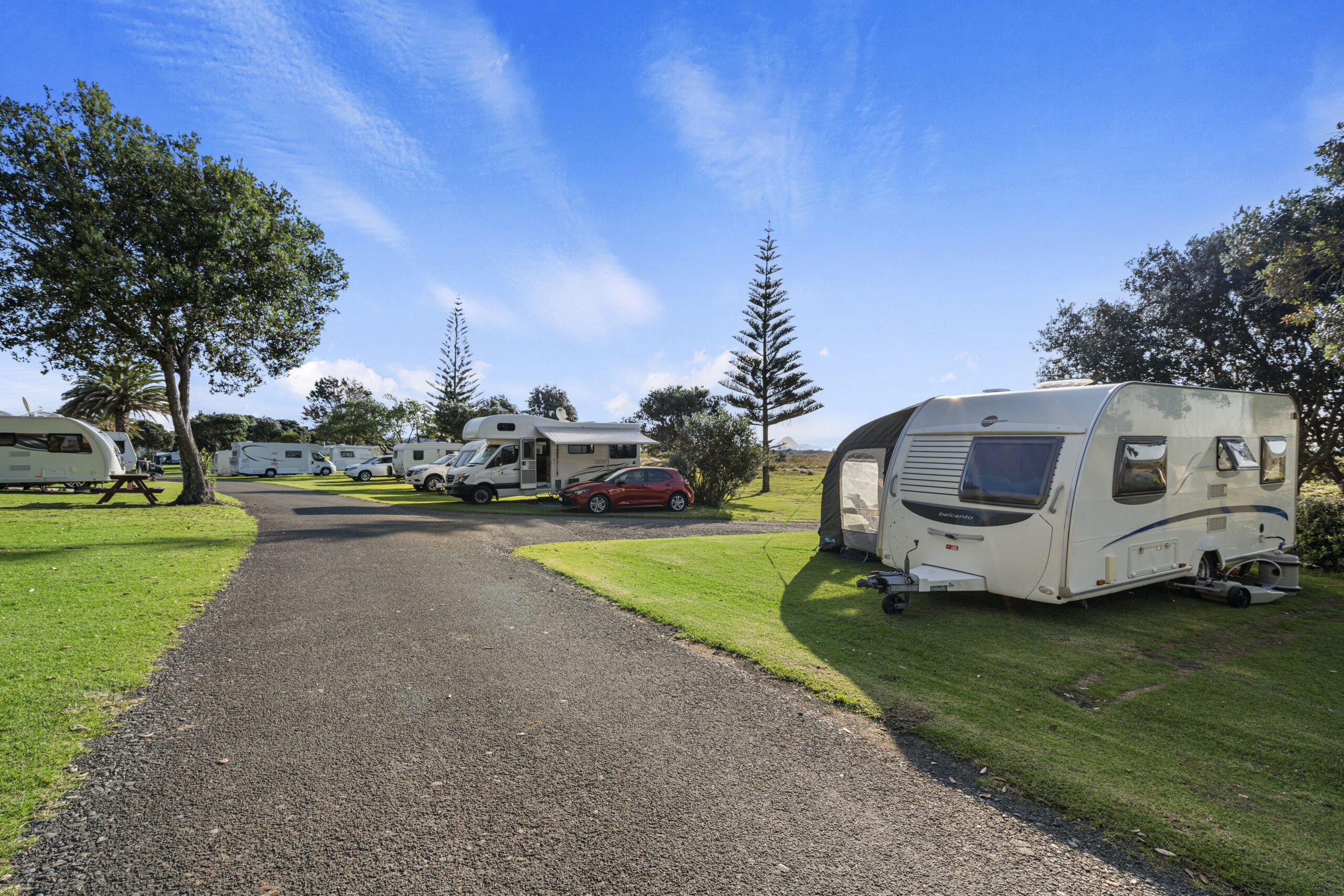 A scenic campground with several caravans and motorhomes parked on grassy plots next to a paved road. Trees and clear blue skies are visible in the background. A red car is parked next to one of the motorhomes. The scene suggests a peaceful outdoor setting.