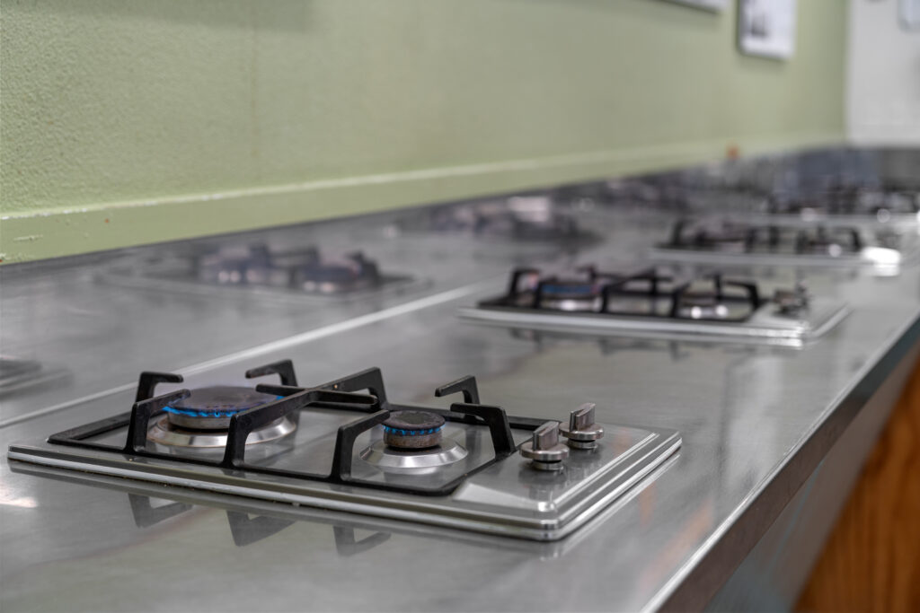 Close-up view of a commercial kitchen's stainless steel countertops featuring several gas burners with blue flames. The burners are aligned in a row against a light green wall, indicating a well-organized cooking space.