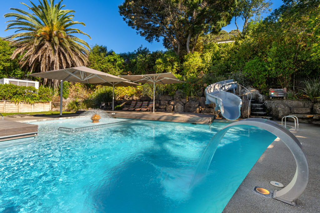 A backyard pool area with a curving waterslide, a fountain, pool chairs, and large umbrellas for shade. Lush greenery, including a tall palm tree, surrounds the pool, and there is a wooden deck adjacent to the water.