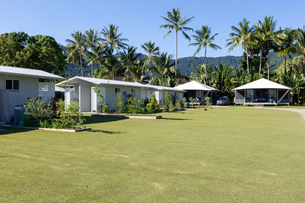 A tropical resort with small white cabins and tent-like structures surrounded by lush greenery and tall palm trees. The foreground features a well-maintained grassy area, and the background has a dense forest with distant hills under a clear blue sky.