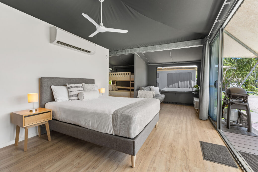 A modern hotel room with a large bed, ceiling fan, and wooden bedside table with lamps. The room also features bunk beds, a couch, and floor-to-ceiling glass doors that open to an outdoor balcony. The decor includes neutral tones and contemporary furnishings.