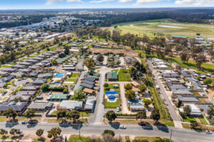 Aerial view of a suburban neighborhood in Bendigo featuring houses with backyards, a swimming pool, and tree-lined streets. In the background, there are green fields and a race track. The area is surrounded by dense greenery and a distant urban landscape.