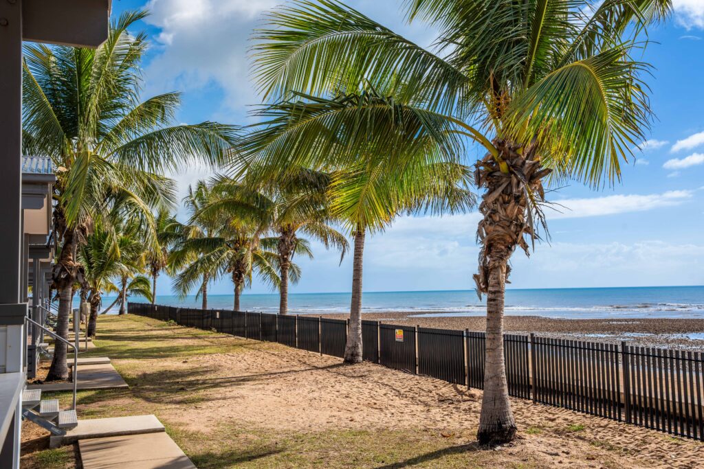 A serene beach scene shows palm trees lining a sandy shore with a black fence separating the beach from nearby buildings. Despite the winter season, the calm ocean stretches out under a blue sky with scattered clouds, creating an unusually tranquil and tropical atmosphere.