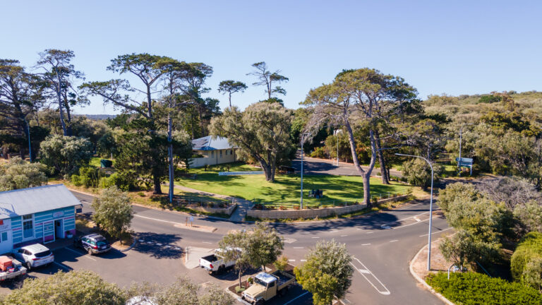 An aerial view of a small roundabout surrounded by lush greenery and trees, capturing the scenic charm of Yallingup. The roundabout provides access to a few streets and parking area with cars. A light blue building is visible on the left side, and a light yellow building sits in the background. The sky is clear and blue.