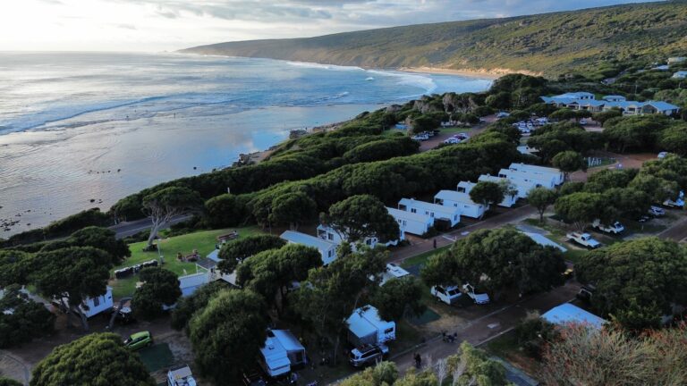 Aerial view of a coastal campsite with white cabins surrounded by lush green trees in Yallingup. The site is near the ocean, with waves gently crashing against the shore, and vibrant wildflowers dotting the landscape. Rolling hills are seen in the background under a partly cloudy sky.