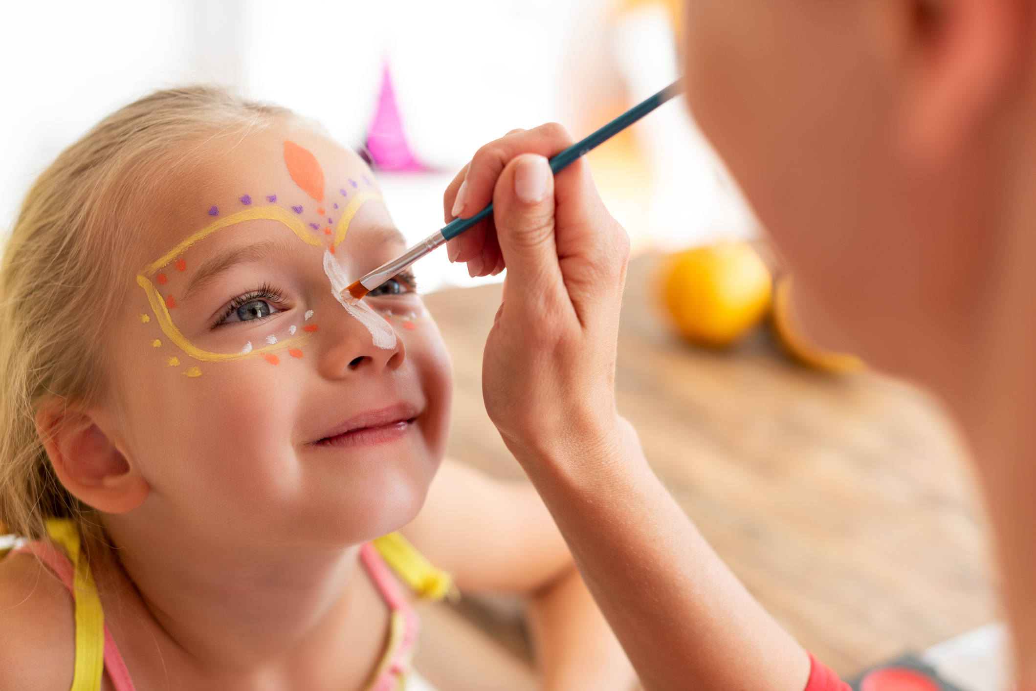 A young girl with a happy expression gets her face painted by an adult. She sits patiently as the adult carefully paints intricate, colorful designs around her eyes and forehead. The background is softly blurred, focusing attention on the painting process.