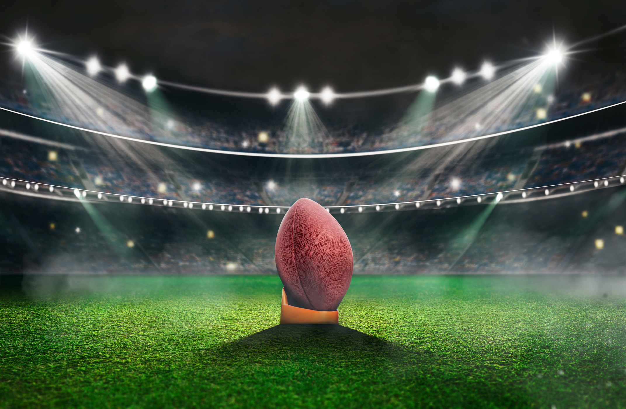 A football stands upright at the center of a brightly lit, empty stadium. Surrounding floodlights illuminate the green turf, and the stadium seating is visible in the background under a dark sky.