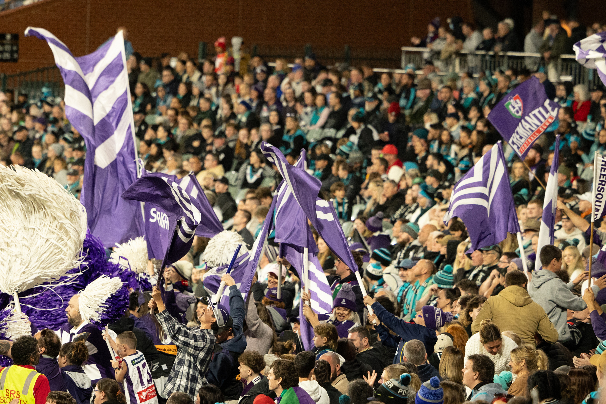 A lively crowd at a sports stadium, many fans waving purple and white flags. The stands are packed with enthusiastic spectators, some dressed in team colors and cheering. The scene is vibrant with movement and energy, indicating a high-stakes, exciting event.