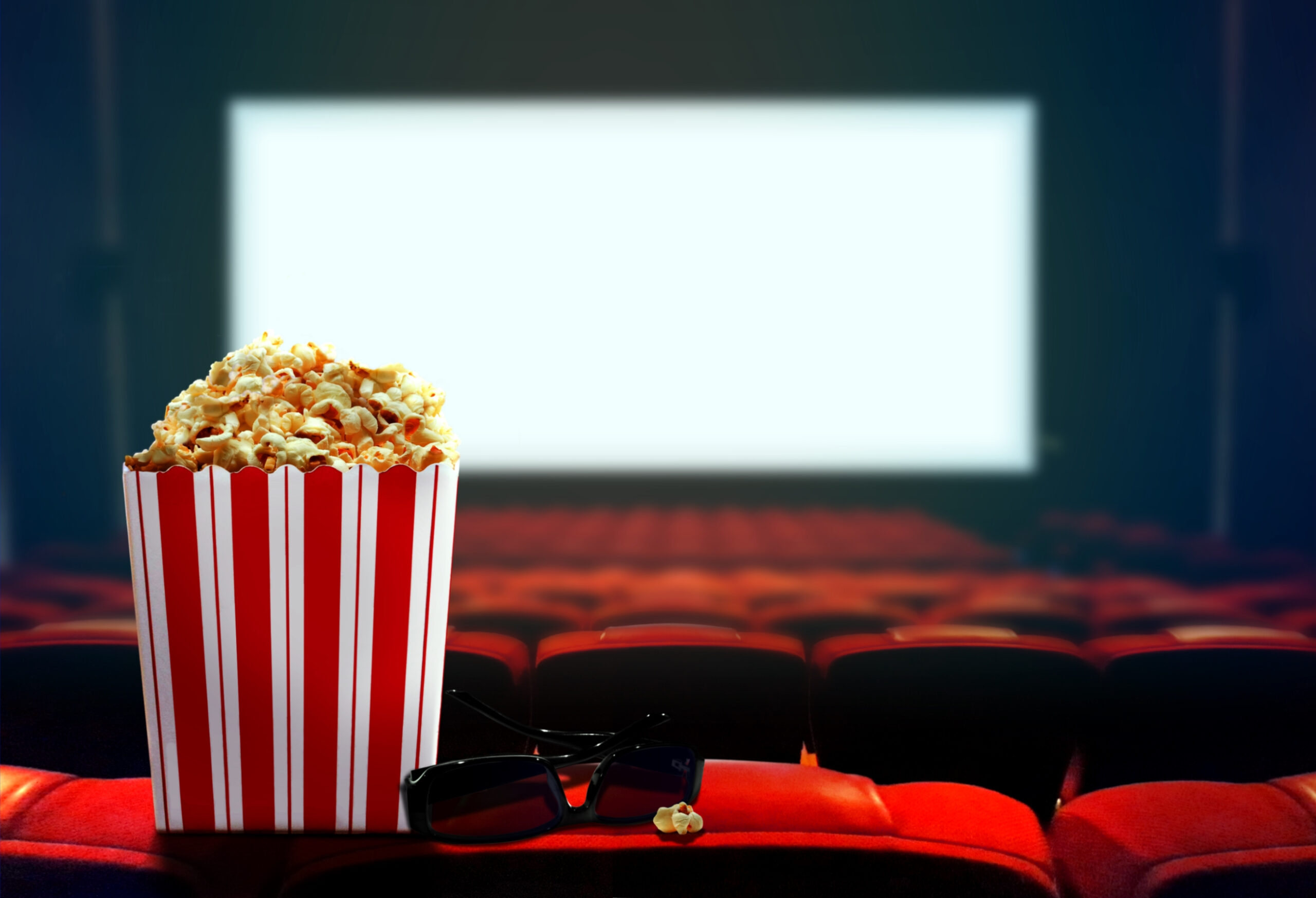 A red and white striped popcorn bucket filled with popcorn sits on a theater seat armrest, next to a pair of black 3D glasses. The background features rows of red seats leading to a bright, blank movie screen. The theater is dimly lit.